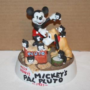 May include: A ceramic figurine of Mickey Mouse and Pluto with a red, white, and blue color scheme. Mickey is holding a small black kitten and Pluto is standing on a dog house with a sign that says "Pluto". The base of the figurine is white and has the text "Walt Disney's Pal Pluto - 1933 - Mickey's"