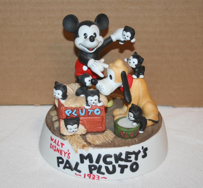 May include: A ceramic figurine of Mickey Mouse and Pluto with a red, white, and blue color scheme. Mickey is holding a small black kitten and Pluto is standing on a dog house with a sign that says "Pluto". The base of the figurine is white and has the text "Walt Disney's Pal Pluto - 1933 - Mickey's"