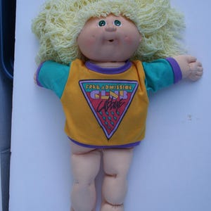 May include: A Cabbage Patch Kid doll with blonde yarn hair wearing a yellow, purple, and teal shirt with the text "Free Admission Club Cabbage" printed on the front.