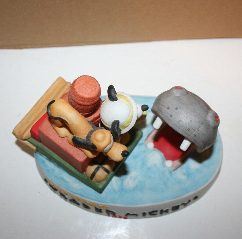 Puede incluir: Una figurita de cer&aacute;mica de Mickey Mouse, Pluto y Goofy en un bote que es perseguido por un hipop&oacute;tamo. La figurita est&aacute; pintada en colores brillantes y tiene un dise&ntilde;o detallado. La base de la figurita est&aacute; inscrita con "Happiest Place on Earth".
