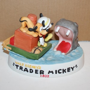 Puede incluir: Una figurita de porcelana de Mickey Mouse y Pluto en un barco con un hipop&oacute;tamo con la boca abierta. La base de la figurita dice "Walt Disney's Trader Mickey 1932".