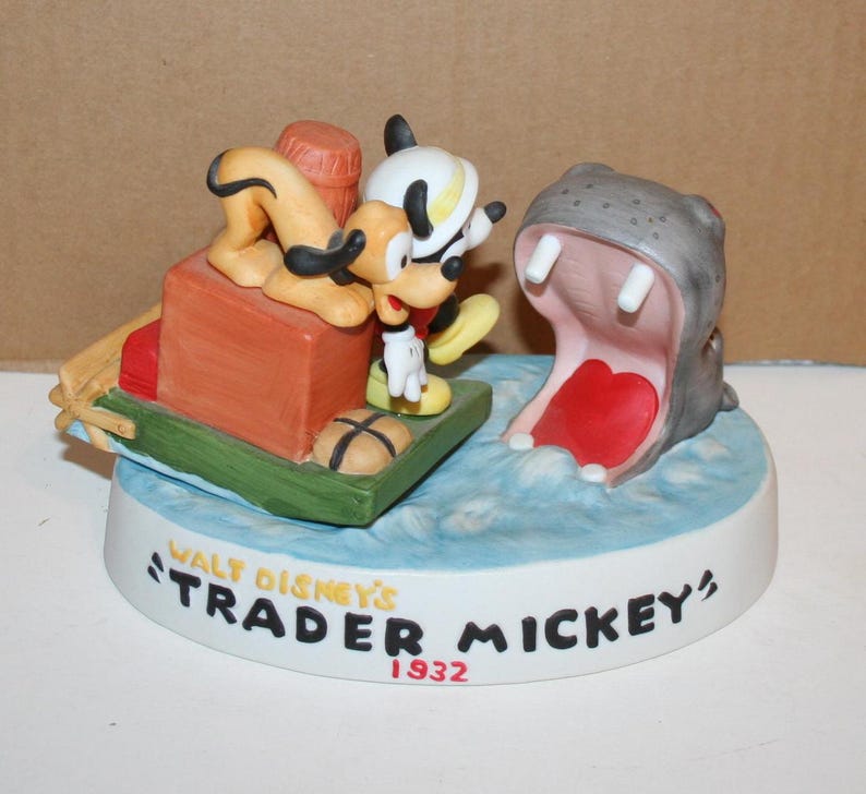 Puede incluir: Una figurita de porcelana de Mickey Mouse y Pluto en un barco con un hipop&oacute;tamo con la boca abierta. La base de la figurita dice "Walt Disney's Trader Mickey 1932".