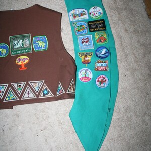 Puede incluir: Un chaleco marr&oacute;n de Girl Scout con varios parches, incluyendo "GLEANERS FOOD BANK" y "Yoga". Una banda de Girl Scout turquesa con varios parches, incluyendo "2009 SS COOKIES", "DETROIT ZOO", "CERAMICS" y "FOOD DRIVE".