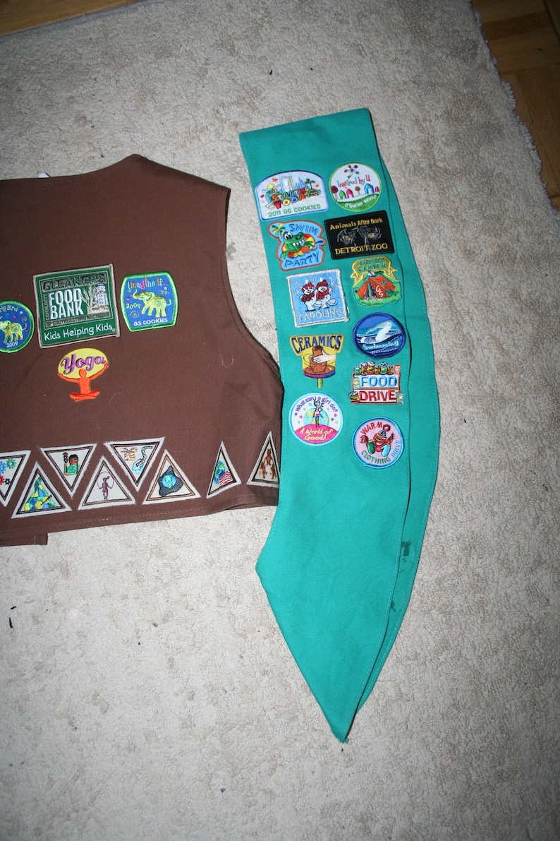 Puede incluir: Un chaleco marr&oacute;n de Girl Scout con varios parches, incluyendo "GLEANERS FOOD BANK" y "Yoga". Una banda de Girl Scout turquesa con varios parches, incluyendo "2009 SS COOKIES", "DETROIT ZOO", "CERAMICS" y "FOOD DRIVE".