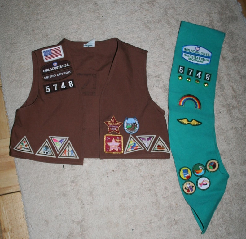 Puede incluir: Chaleco marr&oacute;n de Girl Scout con varios parches e insignias, incluyendo un parche de arco&iacute;ris y un parche con el texto "Girl Scouts USA Southeastern Michigan 5748". Tambi&eacute;n se muestra una faja verde con el texto "Girl Scouts USA Southeastern Michigan 5748" y varios parches.