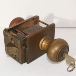 May include: An antique brass door lock with a key inserted. The lock has a rectangular body with a round knob on one side and a smaller, matching knob on the other. The key is silver-colored and attached to a small ring.