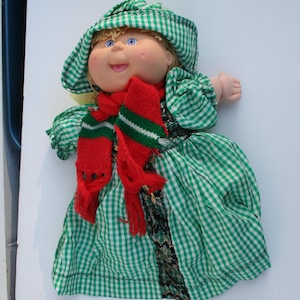 May include: A Cabbage Patch Kid doll wearing a green and white gingham dress, a red and green striped scarf, and a green and white gingham hat.