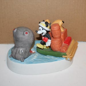 Puede incluir: Una figurita de cer&aacute;mica de Mickey Mouse en un bote con una figurita de hipop&oacute;tamo en el agua. Mickey Mouse lleva una camisa roja y un sombrero amarillo. El bote est&aacute; hecho de madera y tiene un fondo verde. El hipop&oacute;tamo es gris y tiene una boca rosa.