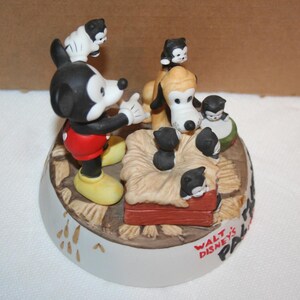 May include: A ceramic figurine of Mickey Mouse and Pluto with a litter of kittens. Mickey is wearing red shorts and yellow shoes. Pluto is a brown and white dog with black ears. The kittens are black and white and are in a basket with straw. The base of the figurine is white with the words "Walt Disney's Pals" in black.