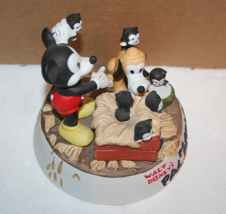 May include: A ceramic figurine of Mickey Mouse and Pluto with a litter of kittens. Mickey is wearing red shorts and yellow shoes. Pluto is a brown and white dog with black ears. The kittens are black and white and are in a basket with straw. The base of the figurine is white with the words "Walt Disney's Pals" in black.
