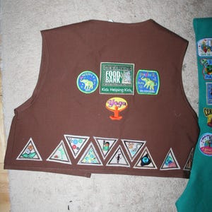 Puede incluir: Chaleco marr&oacute;n de Girl Scout con varios parches, incluyendo "Cleaners Food Bank", "Kids Helping Kids", "Yoga" y "2 SS Cookies". El chaleco tiene una fila de parches triangulares en la parte inferior.