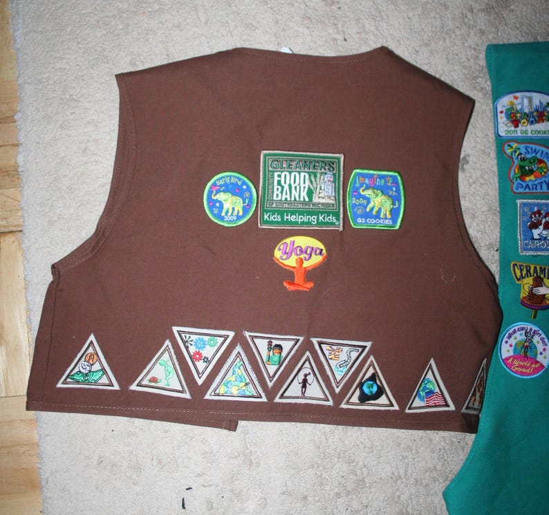 Puede incluir: Chaleco marr&oacute;n de Girl Scout con varios parches, incluyendo "Cleaners Food Bank", "Kids Helping Kids", "Yoga" y "2 SS Cookies". El chaleco tiene una fila de parches triangulares en la parte inferior.