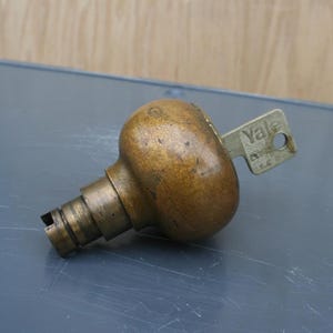 May include: A vintage brass doorknob with a Yale key inserted. The doorknob has a rounded shape and a tarnished finish, showing signs of age. The key is silver with the word "Yale" printed on it. The doorknob is on a gray surface.