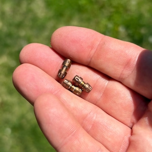 May include: Three small, cylindrical metal beads with a metallic bronze finish. Each bead features a textured, grooved surface with a central band and tapered ends, displaying a distinct industrial design with visible metallic sheen and detailing.