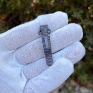 May include: A blue and silver titanium pocket clip with a wavy pattern. The clip is held in a white gloved hand.