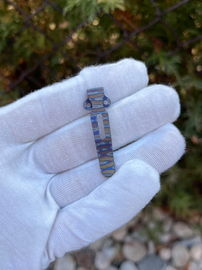 May include: A blue and silver titanium pocket clip with a wavy pattern. The clip is held in a white gloved hand.