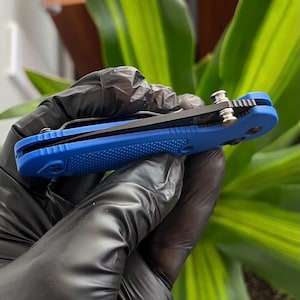 Pode incluir: Uma faca dobrável azul com cabo texturizado e lâmina preta. A faca é segurada em uma mão enluvada, mostrando seu design compacto e detalhes metálicos. A faca tem aproximadamente 7.6 cm de comprimento quando fechada.
