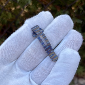 May include: A blue and gray titanium pocket clip with a wavy pattern. The clip is designed to attach to a pocket or belt.