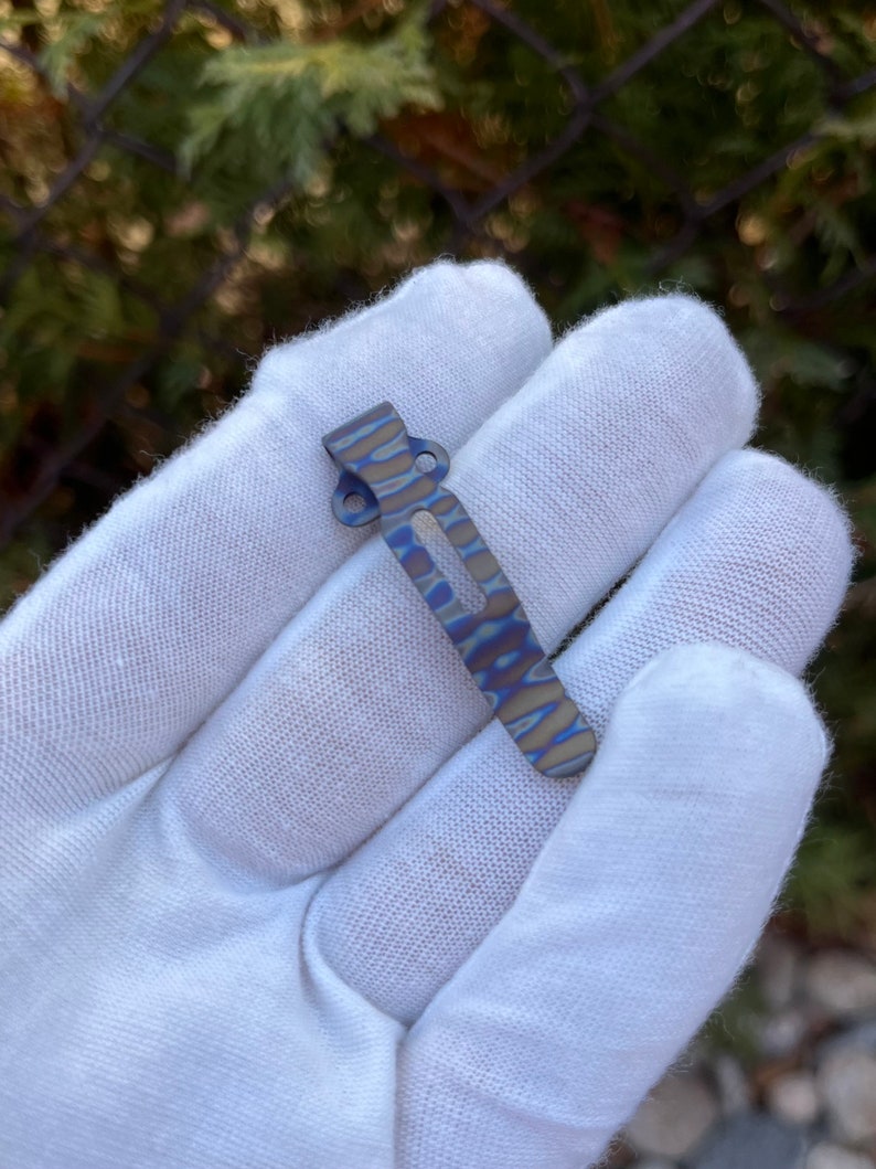 May include: A blue and gray titanium pocket clip with a wavy pattern. The clip is designed to attach to a pocket or belt.