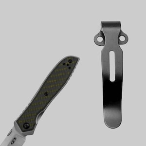 May include: A black pocket clip for a folding knife. The clip has two holes at the top and a slot in the middle.