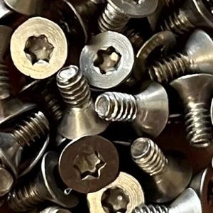 May include: A close-up image of a pile of silver-toned, star-shaped, flat-head screws. The screws have a countersunk head and a hexagonal drive.