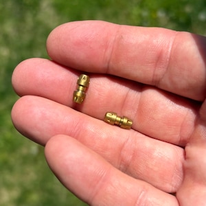 May include: Two small, cylindrical brass components with a metallic gold finish. Each piece features a central narrow shaft flanked by two wider, textured ends with small, evenly spaced indentations around the circumference of the caps.
