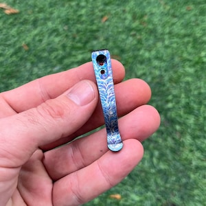 May include: A blue and purple metal pocket clip with a decorative etched pattern. The clip has two holes near the top and a rounded bottom. The clip is held in a hand against a green background.