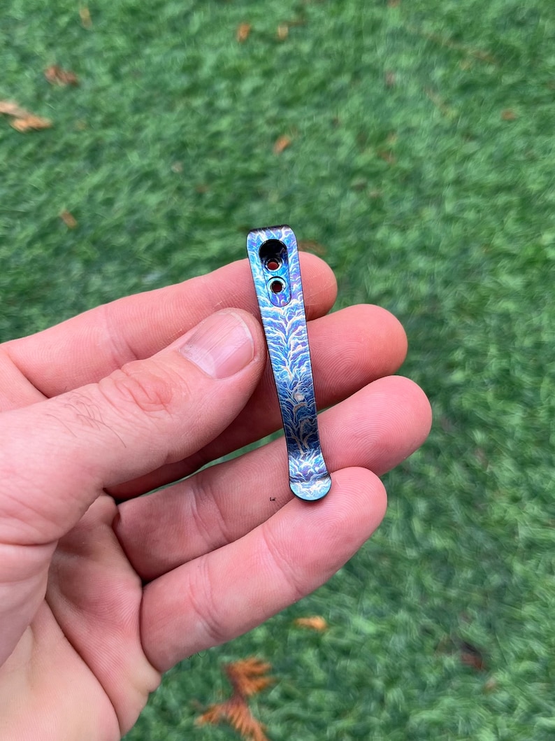 May include: A blue and purple metal pocket clip with a decorative etched pattern. The clip has two holes near the top and a rounded bottom. The clip is held in a hand against a green background.