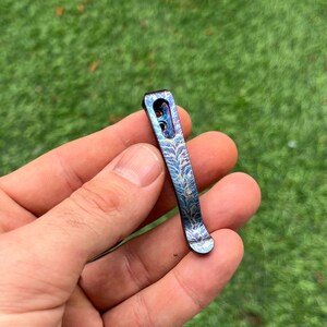 May include: A small, decorative pocket clip with a blue and purple finish. The clip features a leaf-like pattern etched into the metal. The clip is held in a hand against a blurred green background.