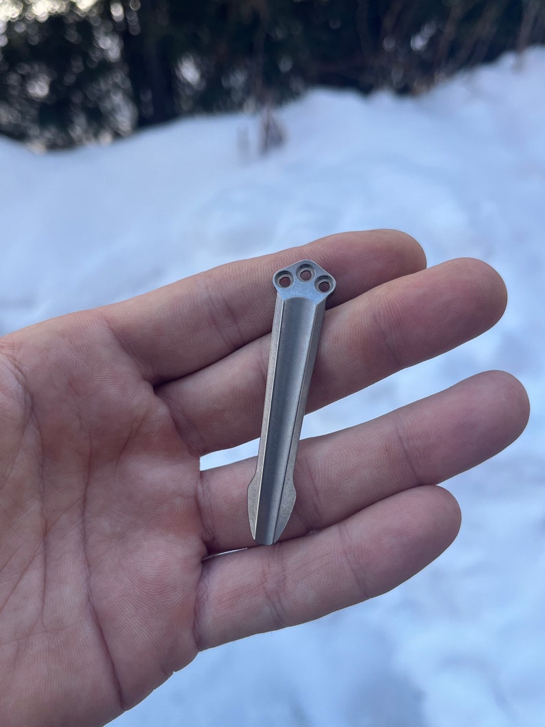 Peut inclure: Un outil en m&eacute;tal argent&eacute; avec une pointe pointue et trois trous en haut. L'outil est tenu dans une main sur un fond enneig&eacute;.