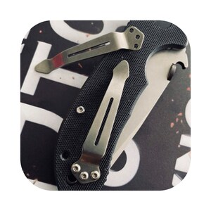 May include: A black pocket knife with a silver clip and a textured handle. The knife is closed and the clip is attached to the handle.