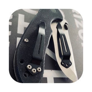May include: A black pocket knife with a textured handle and a silver blade. The knife has a pocket clip on the handle and a small screw on the blade.