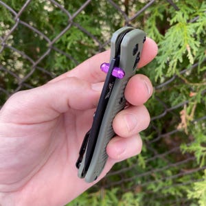 Peut inclure: Un couteau de poche vert et noir avec un accent violet sur la poignée. Le couteau est fermé et a une poignée texturée.