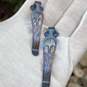 May include: Two blue and purple titanium pocket clip keychains with a decorative design. The keychains are on a white surface.