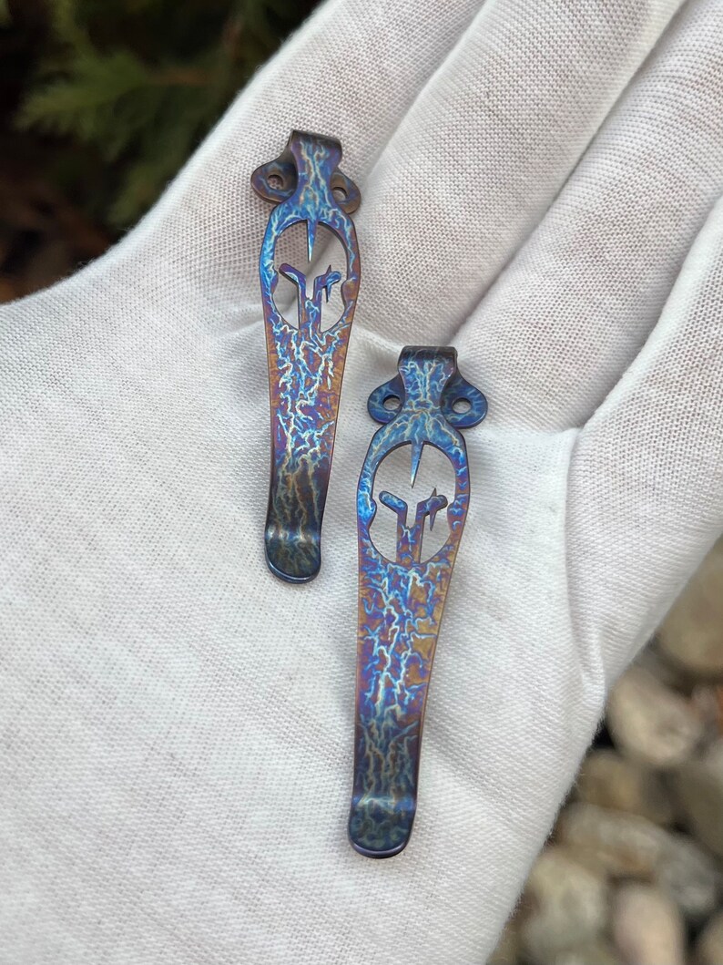 May include: Two blue and purple titanium pocket clip keychains with a decorative design. The keychains are on a white surface.