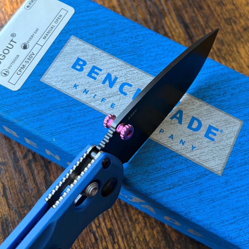 Puede incluir: Un cuchillo Benchmade azul con una hoja negra y detalles rosas. El cuchillo est&aacute; parcialmente abierto, mostrando el filo de la hoja. La caja azul detr&aacute;s del cuchillo tiene texto blanco que dice "Benchmade Knife Company."