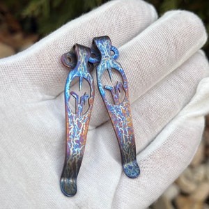 May include: Two blue and purple titanium pocket clip keychains with a unique design. The keychains are made of metal and have a textured finish.