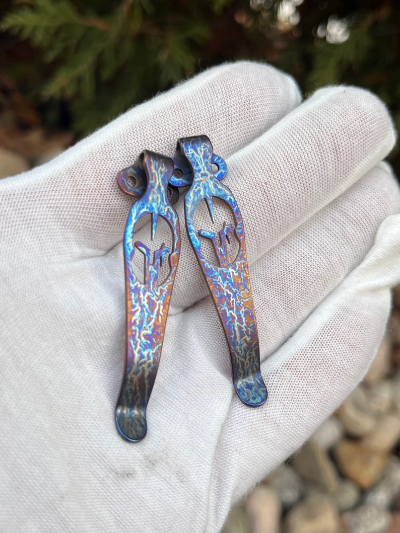 May include: Two blue and purple titanium pocket clip keychains with a unique design. The keychains are made of metal and have a textured finish.