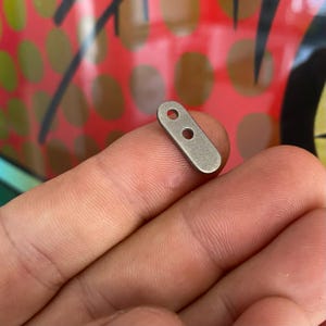 May include: A small, silver-colored metal connector with two holes. The connector is oval-shaped and has a smooth, polished finish.