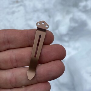 May include: A bronze-colored pocket clip with a rectangular body and a pointed end. The clip has a long, narrow slot down the center and three small holes at the top. The clip is designed to attach to a pocket or other item.