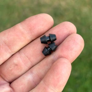 Può includere: Due piccoli componenti metallici cilindrici neri con filettatura esterna su entrambe le estremità e una sezione centrale liscia, stretta e non filettata. Questi elementi di ferramenta presentano una distinta texture scanalata sulle aree filettate.