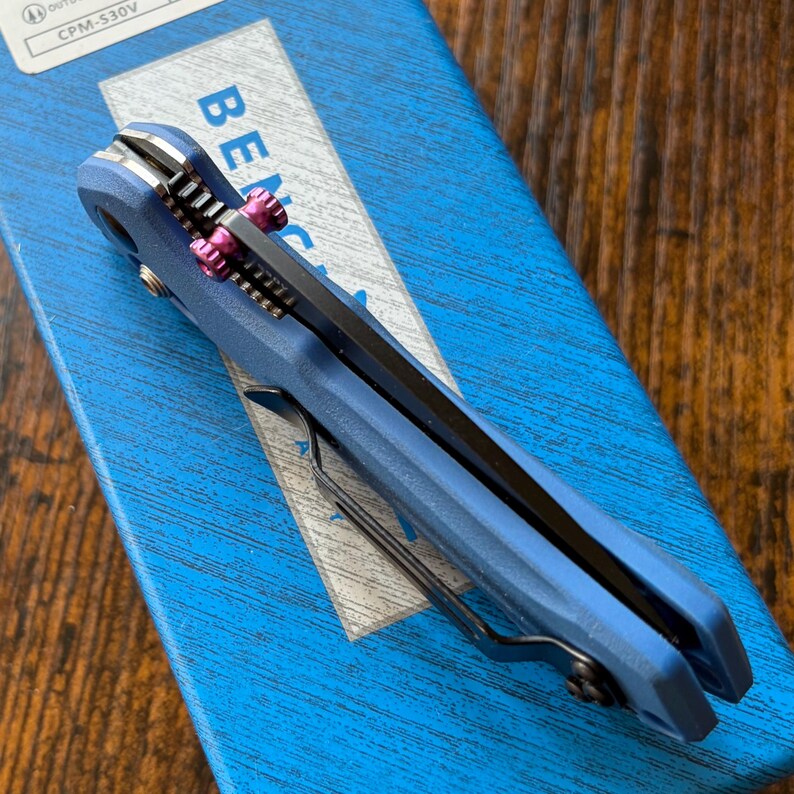 Puede incluir: Una navaja plegable azul con una hoja negra y detalles rosas. El cuchillo est&aacute; sobre una caja azul. El cuchillo tiene un clip para el bolsillo y un mango texturizado. La palabra "Benchmade" es visible.