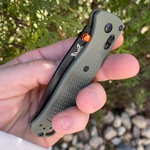 Puede incluir: Un cuchillo plegable verde con una hoja negra y un acento naranja en el mango. El cuchillo tiene un mango texturizado y un logotipo de mariposa en la hoja.