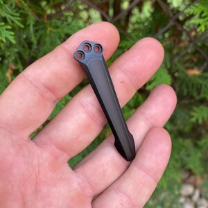 May include: A black metal pocket clip with three circular holes at the top. The clip is rectangular with a curved end. The background is a blurred view of green foliage.