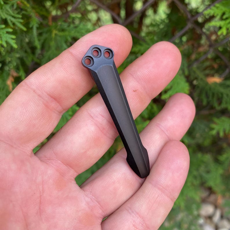 May include: A black metal pocket clip with three circular holes at the top. The clip is rectangular with a curved end. The background is a blurred view of green foliage.
