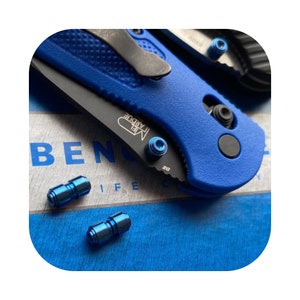 Puede incluir: Un cuchillo de bolsillo azul y negro con un mango azul y una hoja negra. El cuchillo está abierto y la hoja es visible. Hay tres tornillos azules en el cuchillo. El cuchillo está sobre una superficie azul y blanca.