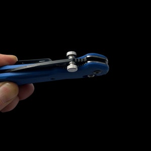 Puede incluir: Un cuchillo de bolsillo azul con una hoja negra y un mecanismo plateado. El cuchillo está abierto y la hoja está parcialmente extendida.