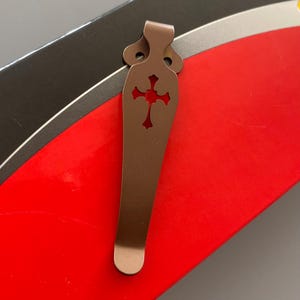May include: A brown metal pocket clip with a cross-shaped cutout. The clip is designed to attach to a pocket or belt.