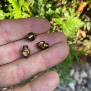 Puede incluir: Primer plano de tres objetos metálicos de color bronce, con una base roscada y una parte superior puntiaguda y facetada. Estos pequeños componentes de hardware se sostienen en una mano, sobre un fondo verde borroso. Probablemente pequeños tornillos.