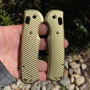 May include: Two gold-colored knife scales with a textured pattern. The scales have multiple holes and are made of metal.